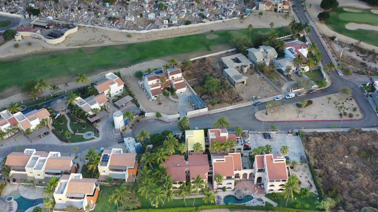 Drone view over homes, pools, parked cars, dry lots, and green fairway area.