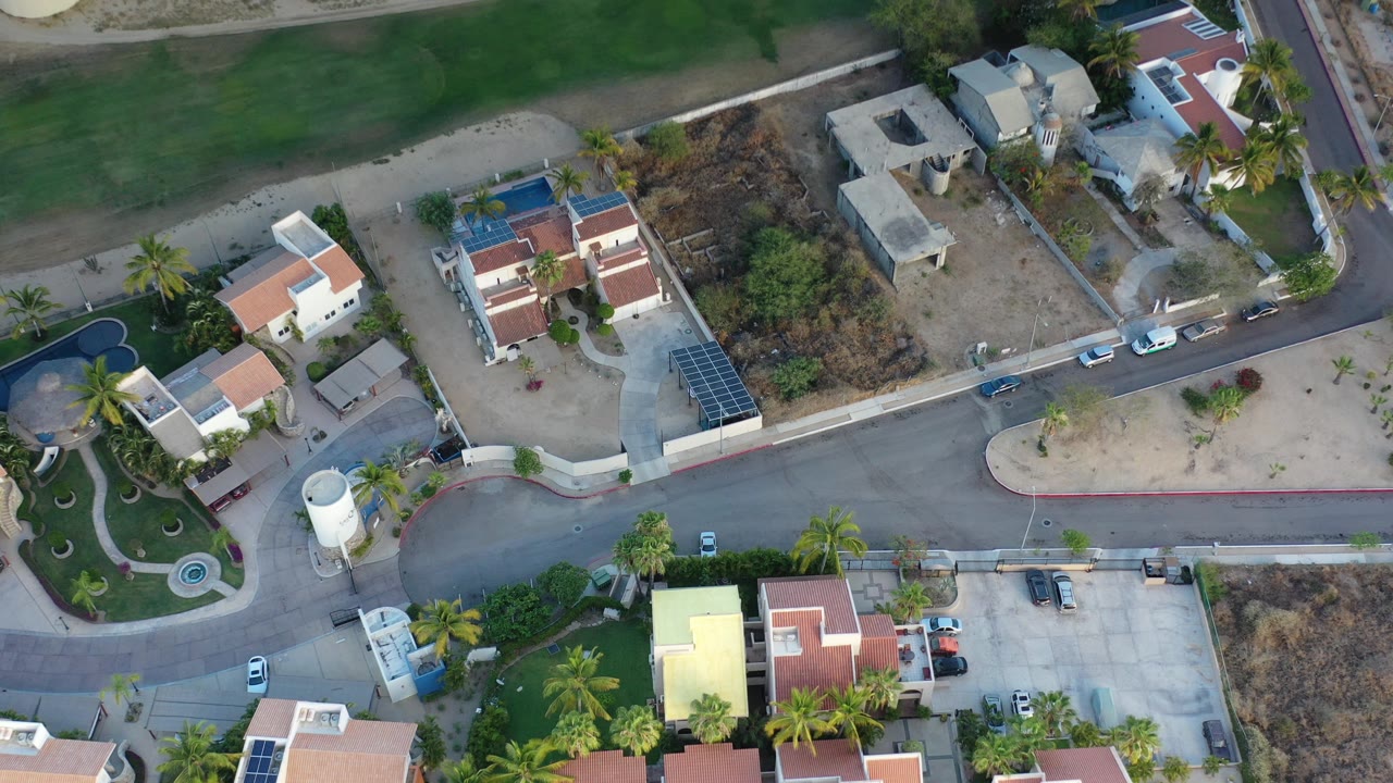 Overhead drone frame in Los Cabos showing white homes with terracotta roofs, green lawns, palm trees, curved roads, small pools, sandy lots, parked cars, and no clearly visible people.