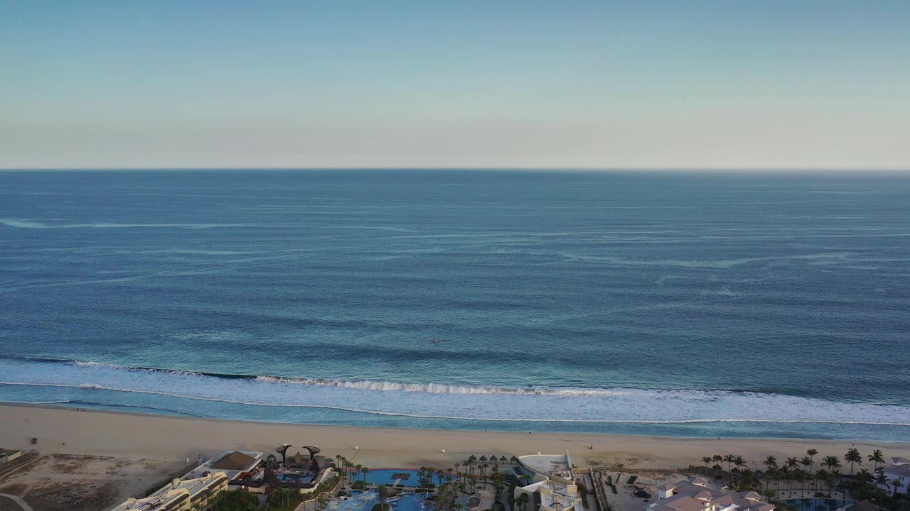 Opening aerial frame over the Los Cabos coast with calm blue water, a straight horizon, white surf bands, tan sand, and no visible people.