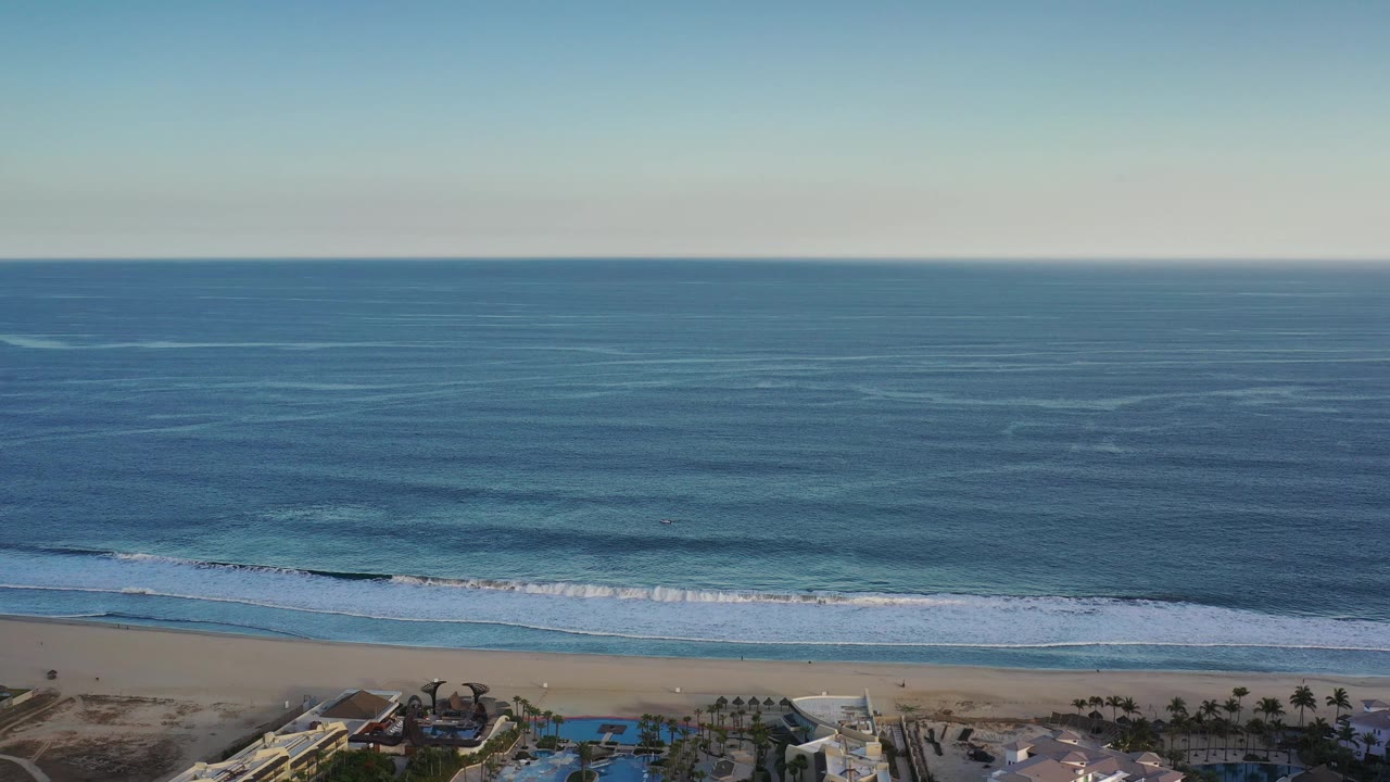 Aerial view in Los Cabos with the horizon high above layered blue water, a thin surf line near shore, beachfront pools at the lower edge, and no visible people.