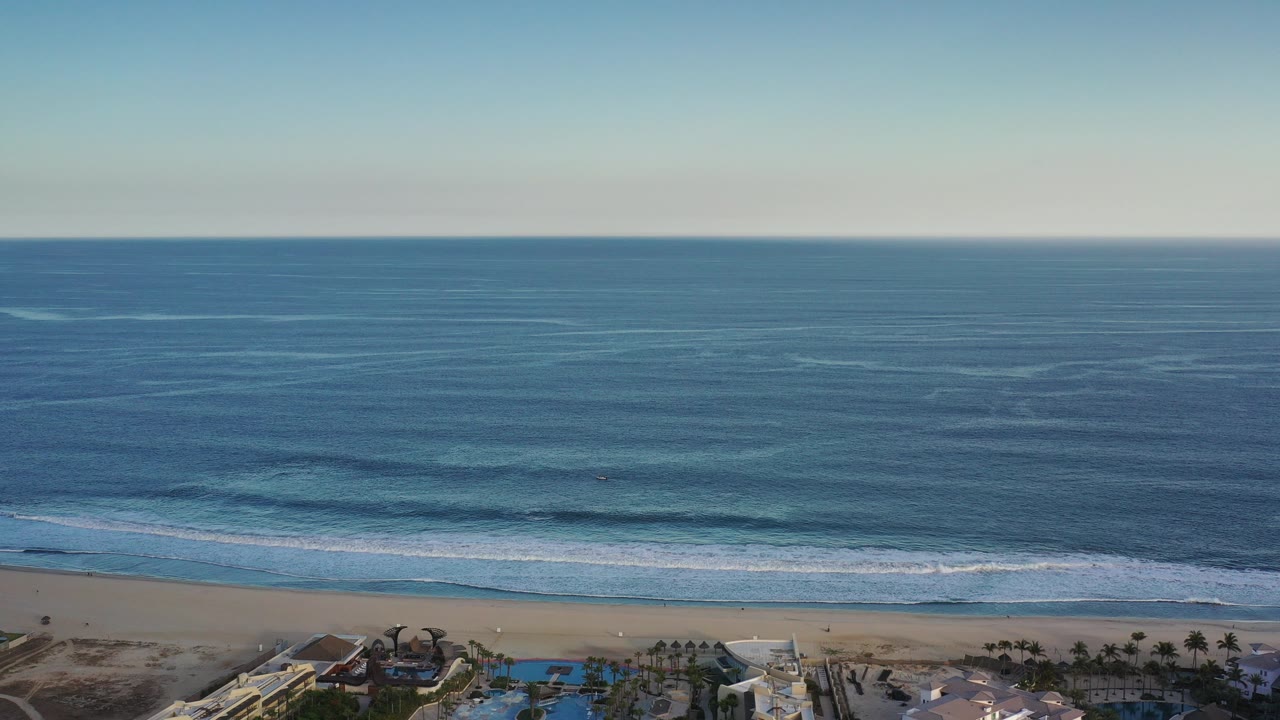 Opening aerial view in Los Cabos, Mexico, with blue ocean across the frame, white surf nearing the sand, beachfront rooftops and pools at the bottom, and no visible people.
