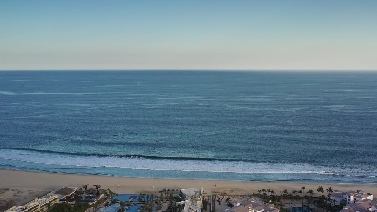 Aerial frame over the Los Cabos coastline showing open blue ocean, a thin surf line, pale sand, and resort rooftops with pools along the lower edge, with no visible people.