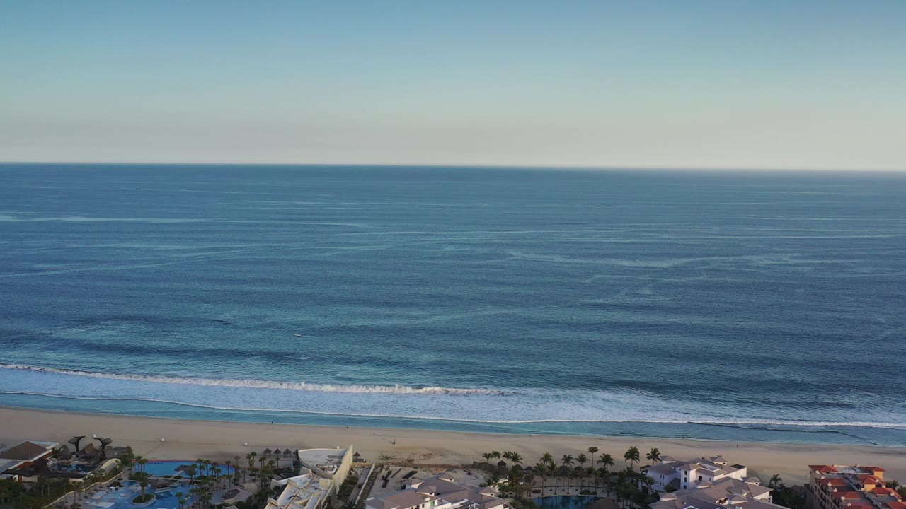 Aerial view of blue ocean waves reaching a sandy beach with buildings at the bottom edge.