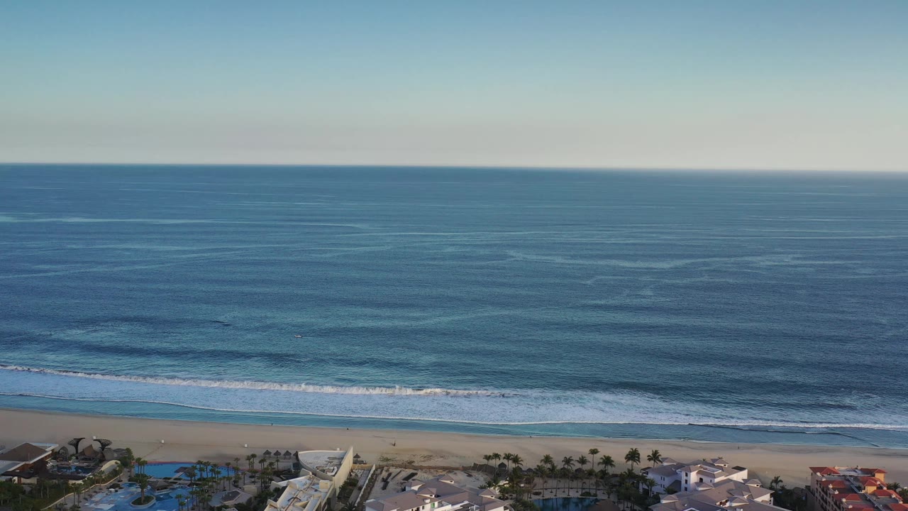 Los Cabos aerial shoreline view with wave bands sliding toward the beach, low rooftops along the bottom edge, and no visible people.