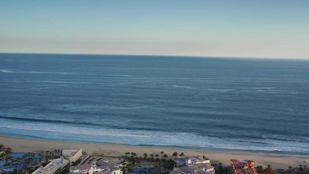 Opening frame of a Los Cabos oceanfront from above, with blue water, a pale clear sky, surf lines, beachfront buildings at the bottom edge, and no visible people.