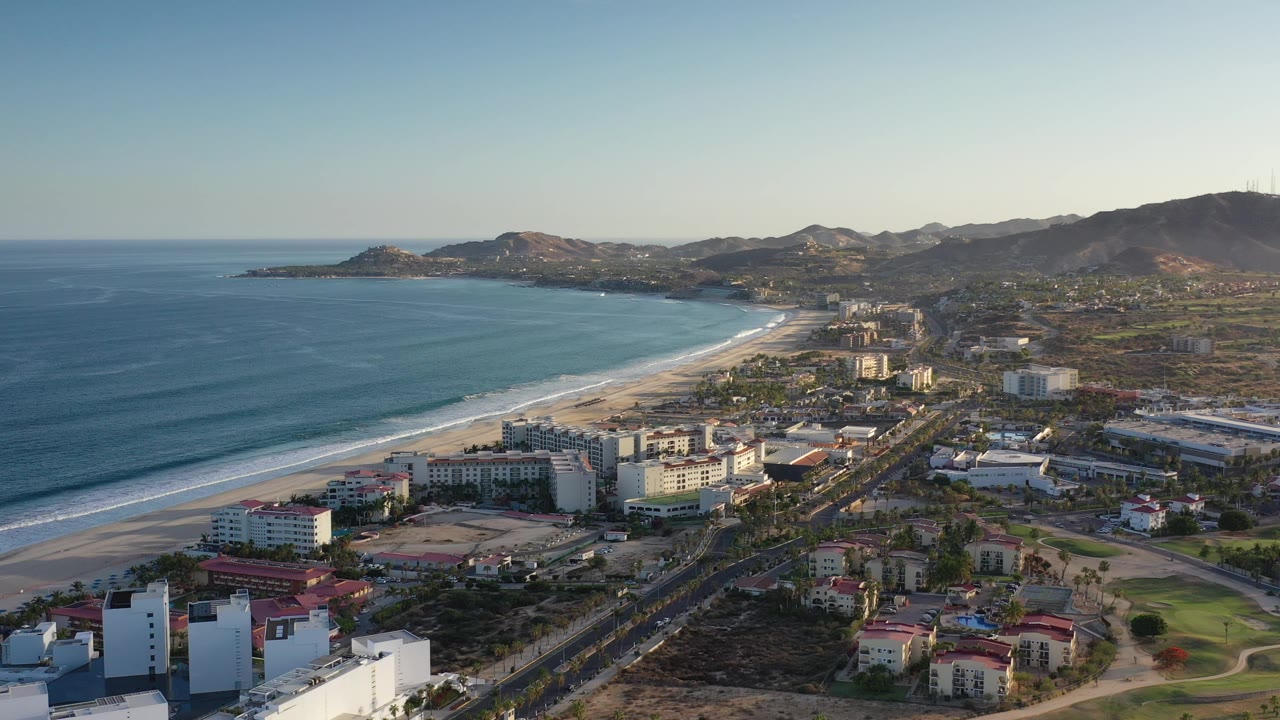 Aerial frame over the Los Cabos coastline with blue ocean at left, a curving beach, beachfront buildings in the foreground, hills beyond the town, and no visible people.