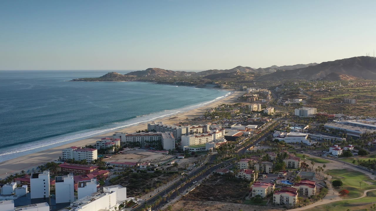 Aerial view in Los Cabos looking along the shoreline, with white surf bands reaching the beach, hotel-like buildings near the sand, inland roads, and no visible people.