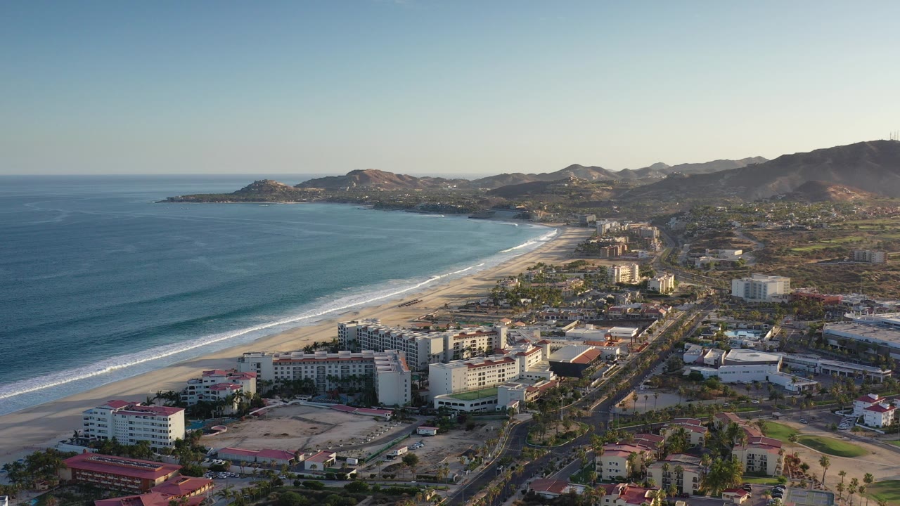 Aerial view of ocean, beach, resort buildings, roads, and mountains.