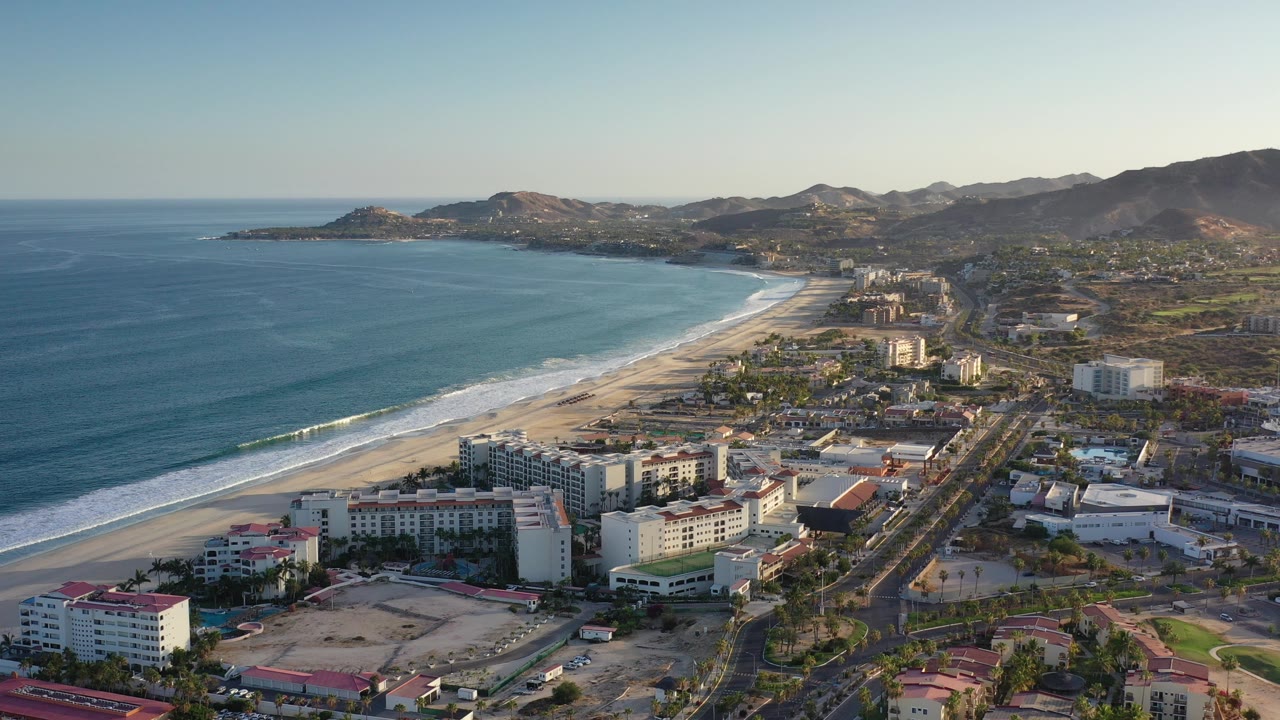A wide aerial frame over Los Cabos, Mexico shows blue ocean, a curving sandy beach, beachfront hotel buildings, inland roads, and dry hills in warm light, with no visible people.