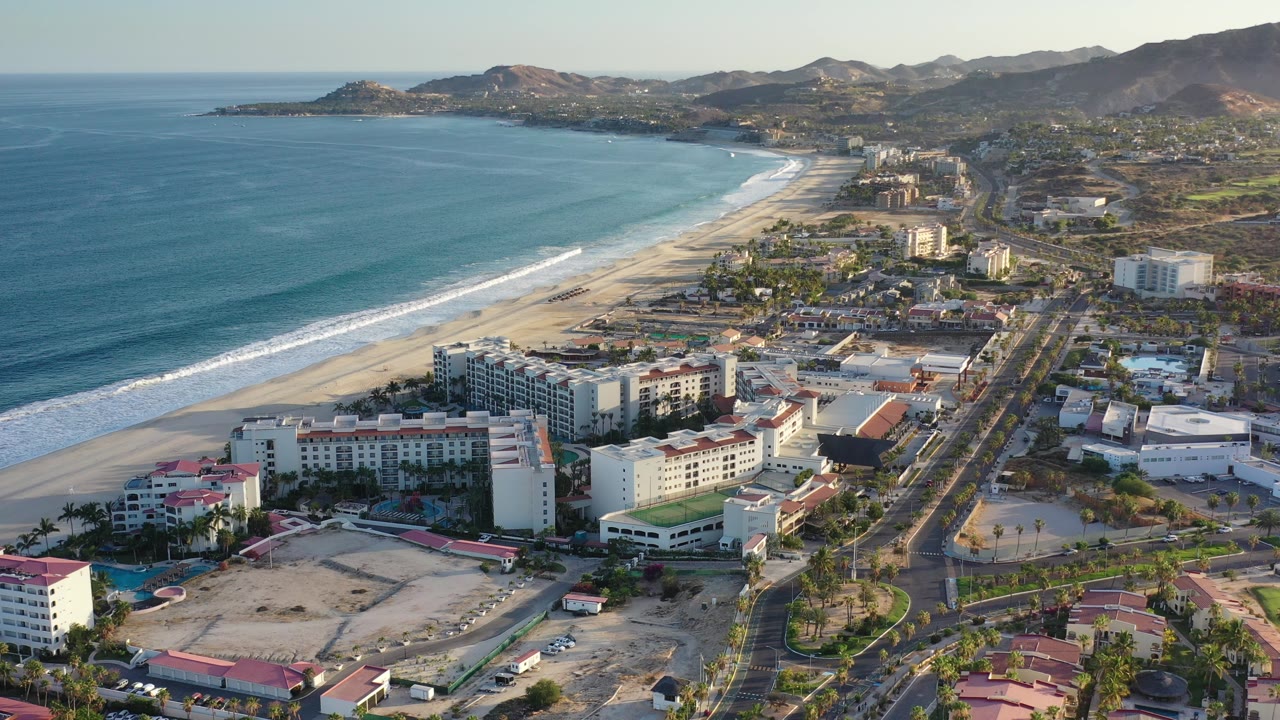 Opening aerial frame over Los Cabos, Mexico, with turquoise ocean at left, a long beach, beachfront hotels, roads, dry hills, distant mountains, and no visible people.