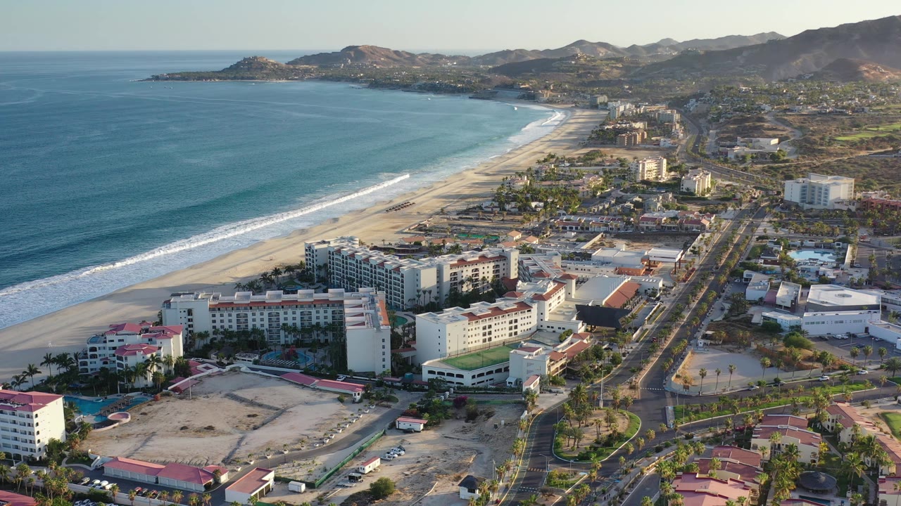 Aerial view in Los Cabos, Mexico, slightly farther along the same shoreline, with white surf bands, hotel blocks, a road grid, hills, mountains, and no visible people.