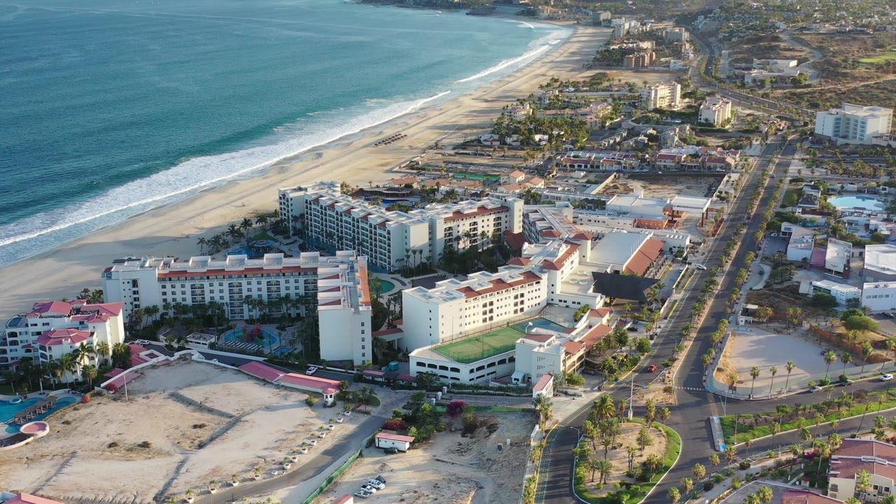 A high aerial frame over Los Cabos shows blue ocean surf, a wide sandy beach, beachfront hotel buildings, pools, and a straight road with no visible people.