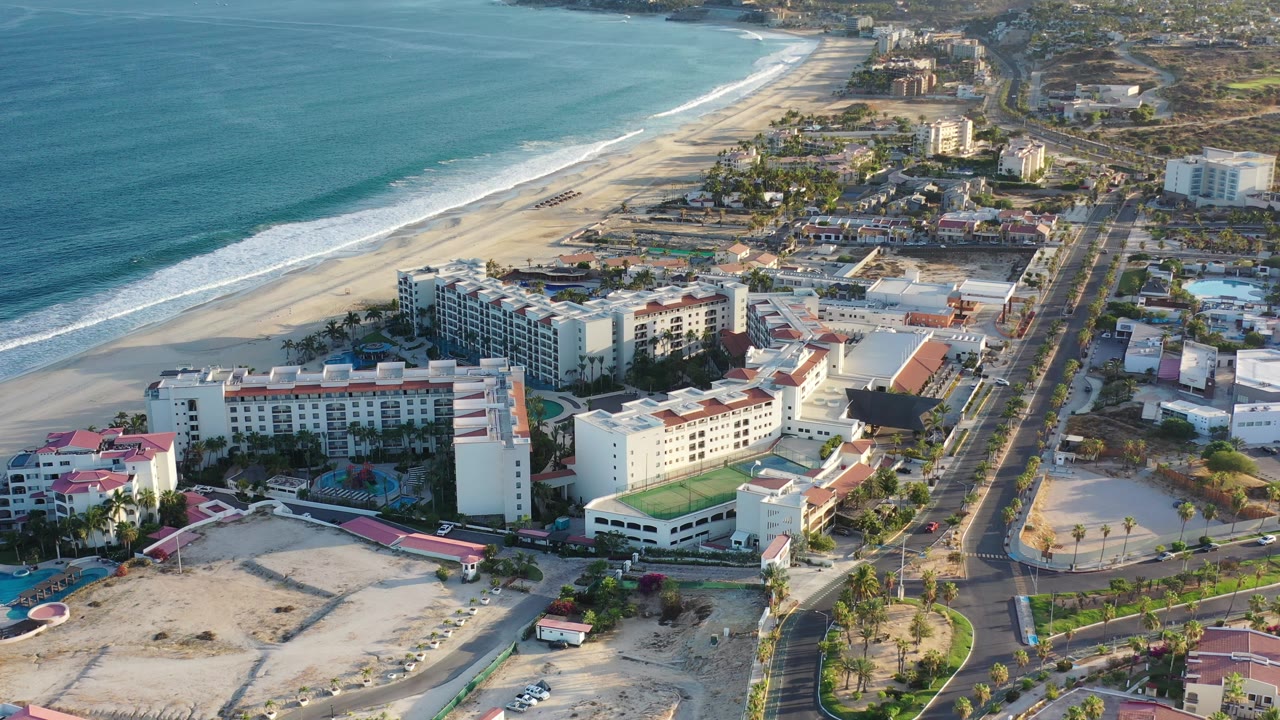 At 0:01, the Los Cabos aerial view keeps the surf and beach along the left while hotel blocks, pools, palms, and the parallel road fill the center and right with no visible people.