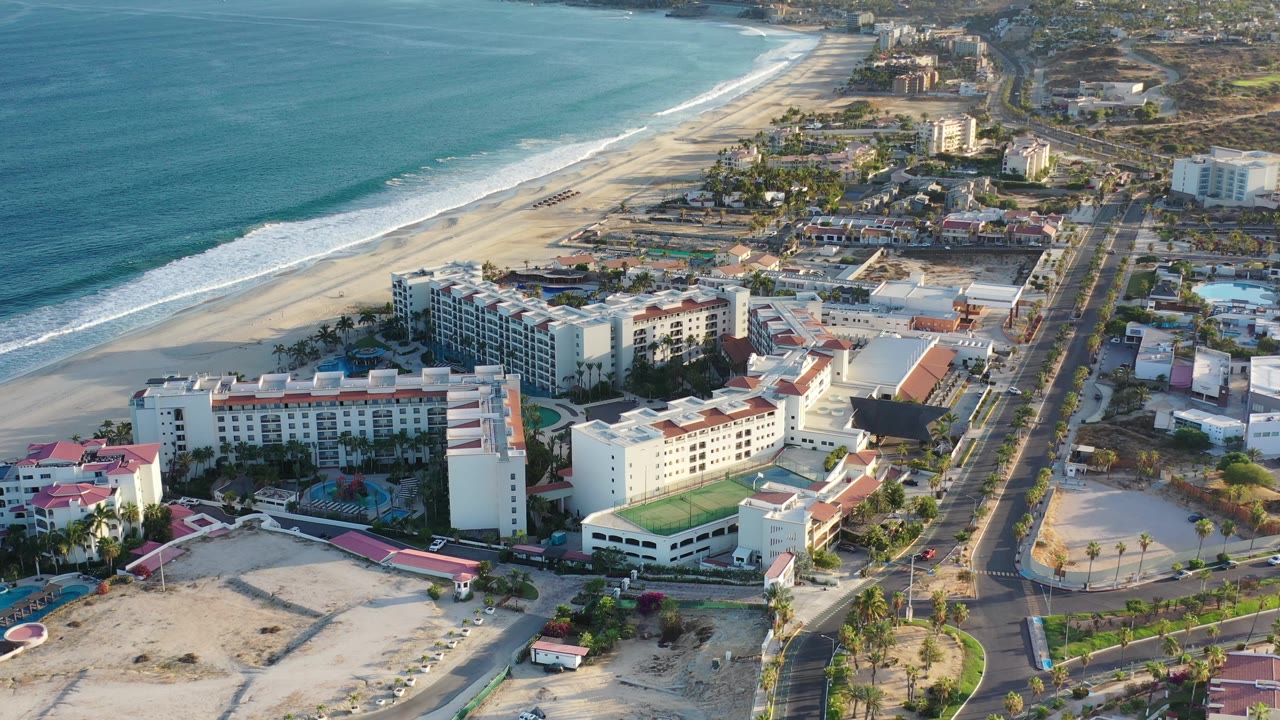 Drone view of Los Cabos shoreline with hotels, pools, and roadways.