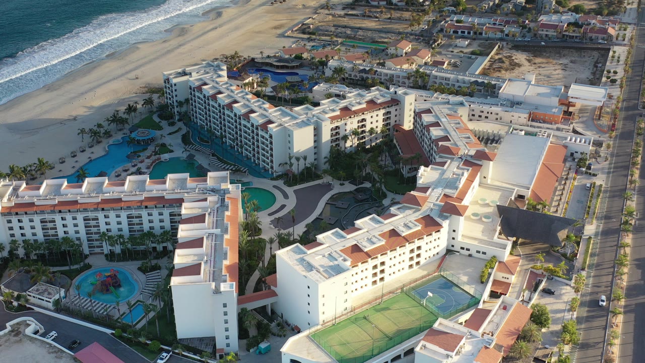 Aerial view over a Los Cabos resort with white buildings, red tile roofs, turquoise pools, tennis courts, a roadside edge, beach, and ocean surf, with no visible people.