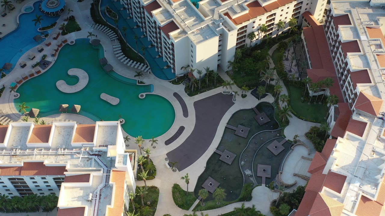 Aerial view in Los Cabos, Mexico, looking down on turquoise curved pools, white buildings, terracotta roof sections, walkways, palm landscaping, and no visible people.