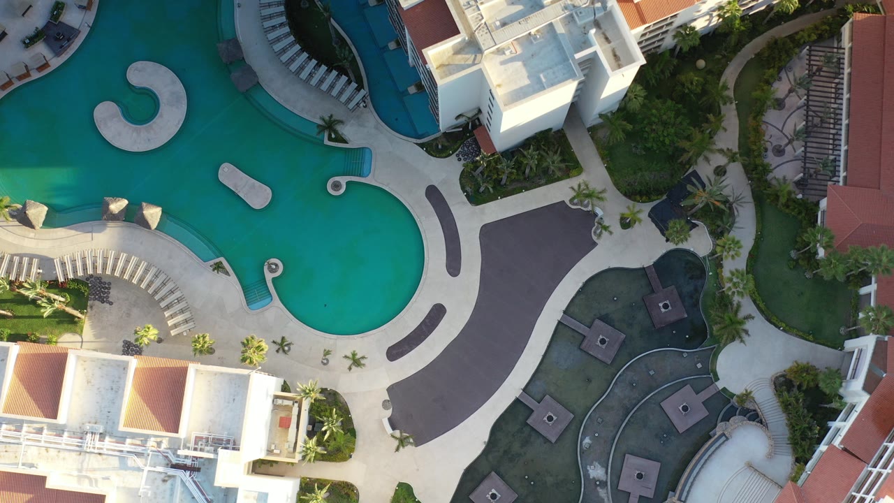 Overhead view in Los Cabos, Mexico, showing curved turquoise resort pools, pale walkways, white buildings, palm landscaping, and no visible people.
