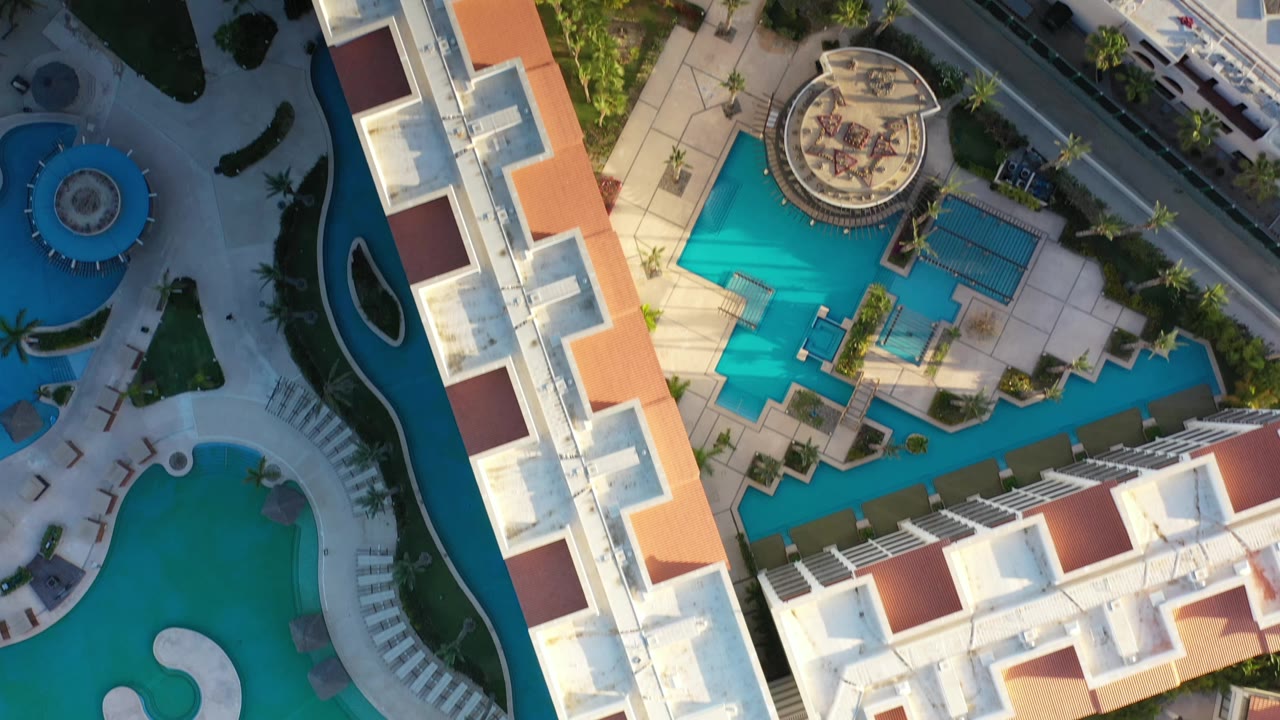 Top-down Los Cabos resort view with a rectangular white building edge, bright blue pool courtyards, curved water features, palm trees, and no visible people.