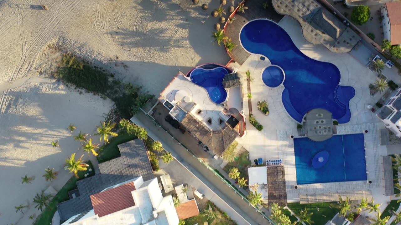 A second overhead Los Cabos frame keeps the large blue pool centered with beach sand to the left, palm landscaping below, and no visible people.