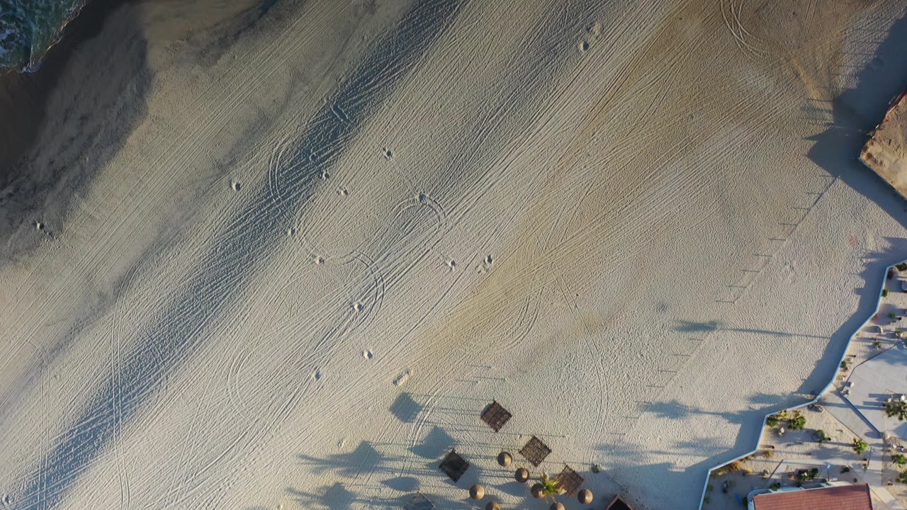 Top-down Los Cabos beach frame with diagonal sand ridges, scattered footprints, a dark wet-sand band near the water, small structures at the lower edge, and no visible people.