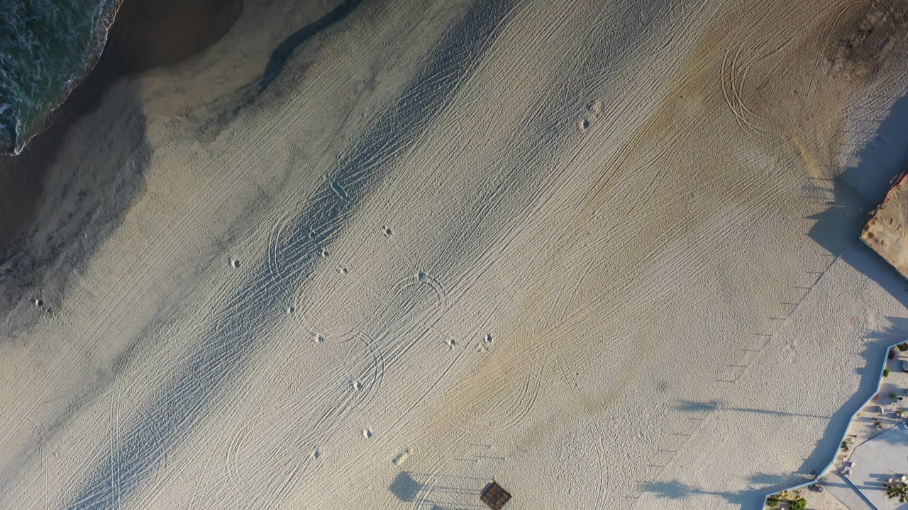 Aerial sand patterns with footprints, long shadows, and a narrow strip of surf near the top right.
