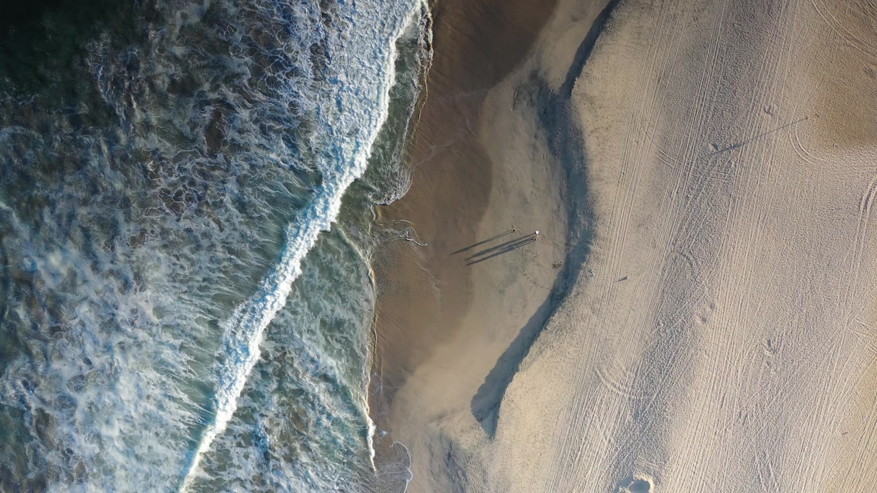 Overhead view in Los Cabos, Mexico, showing white surf along the left edge, pale sand on the right, a dark wet channel, beach tracks, and tiny distant people.
