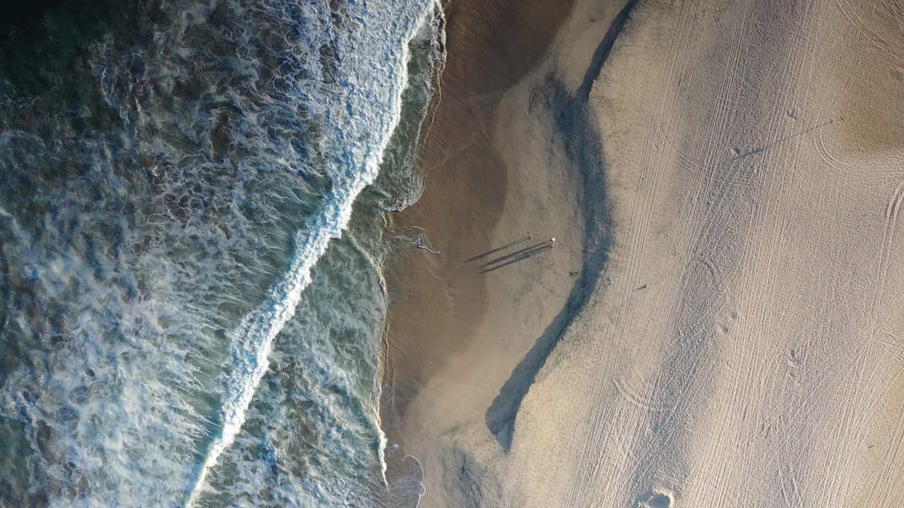 Overhead Los Cabos beach frame with foamy water pushing farther across the left side, smooth tan sand to the right, a curving wet channel, and tiny visible people near the channel.