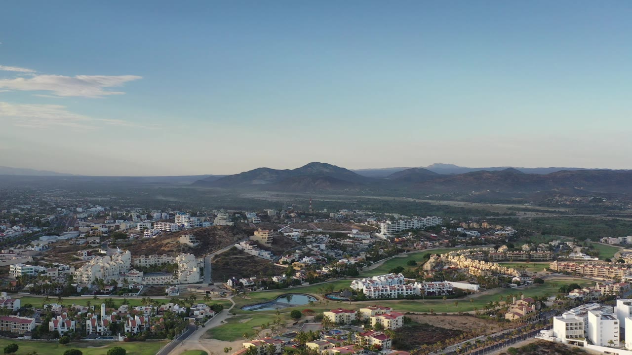 Opening aerial frame over Los Cabos, Mexico, with white buildings and green fairways in the foreground, dry hills beyond, mountains on the horizon, and no visible people.