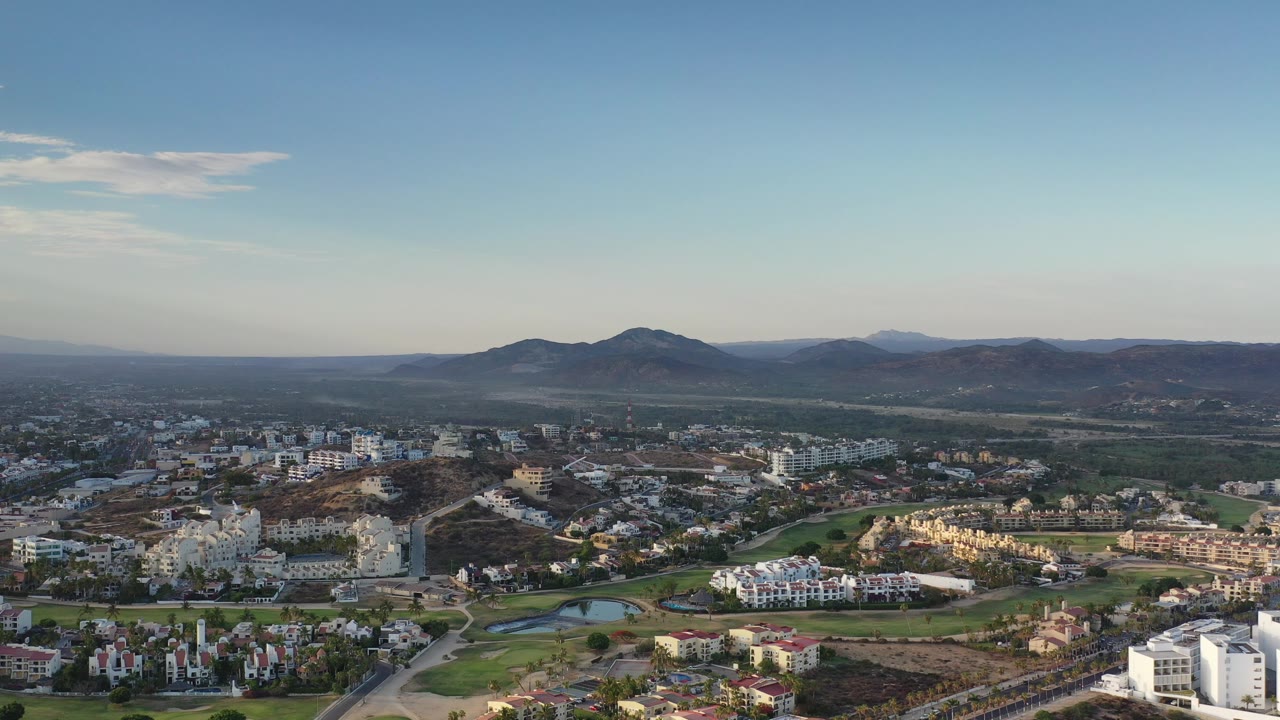Wide aerial landscape with Los Cabos buildings, fairways, hills, mountains, and clear sky.