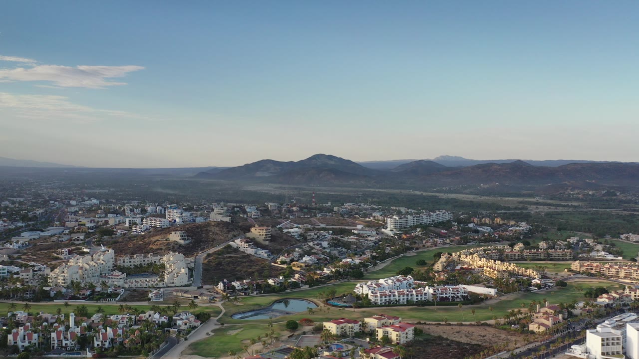 Los Cabos, Mexico aerial frame looking across white residential buildings, a green golf course, small ponds, dry terrain, and distant hills under clear sky, with no visible people.