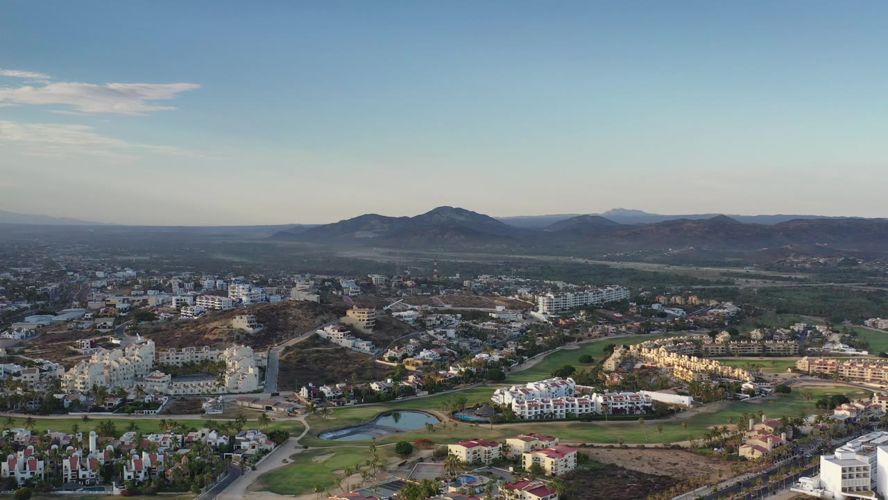 Drone-height view across villas, roads, golf greens, scrubland, and rounded hills.