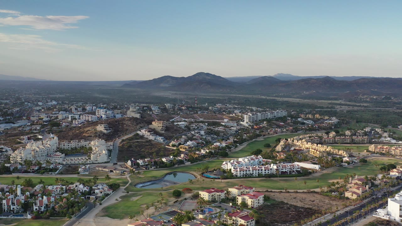 Aerial view over Los Cabos neighborhoods, green fairways, ponds, hills, and distant mountains.