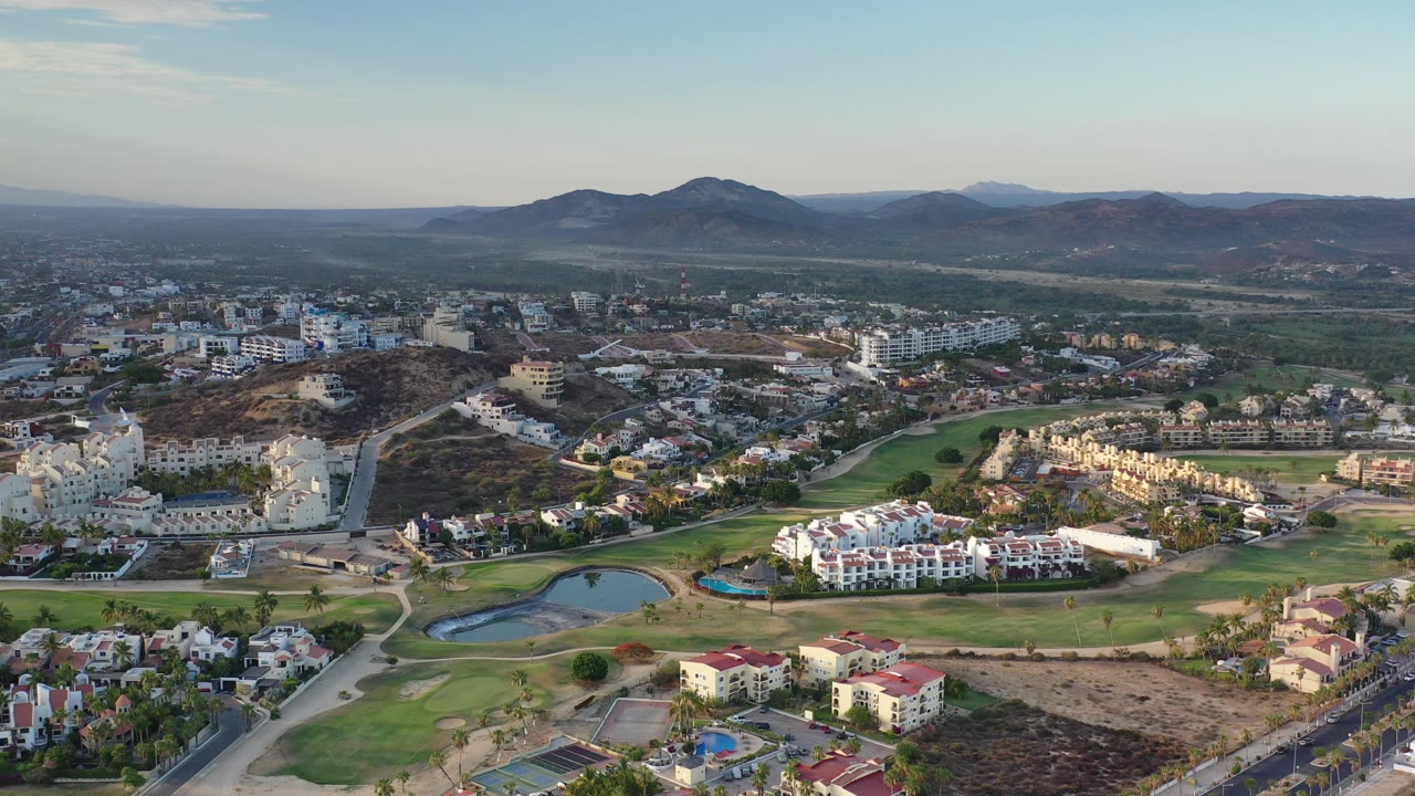 Wide aerial view in Los Cabos, Mexico, showing residential buildings, golf course greens, small ponds, dry hills, and distant mountains with no visible people.