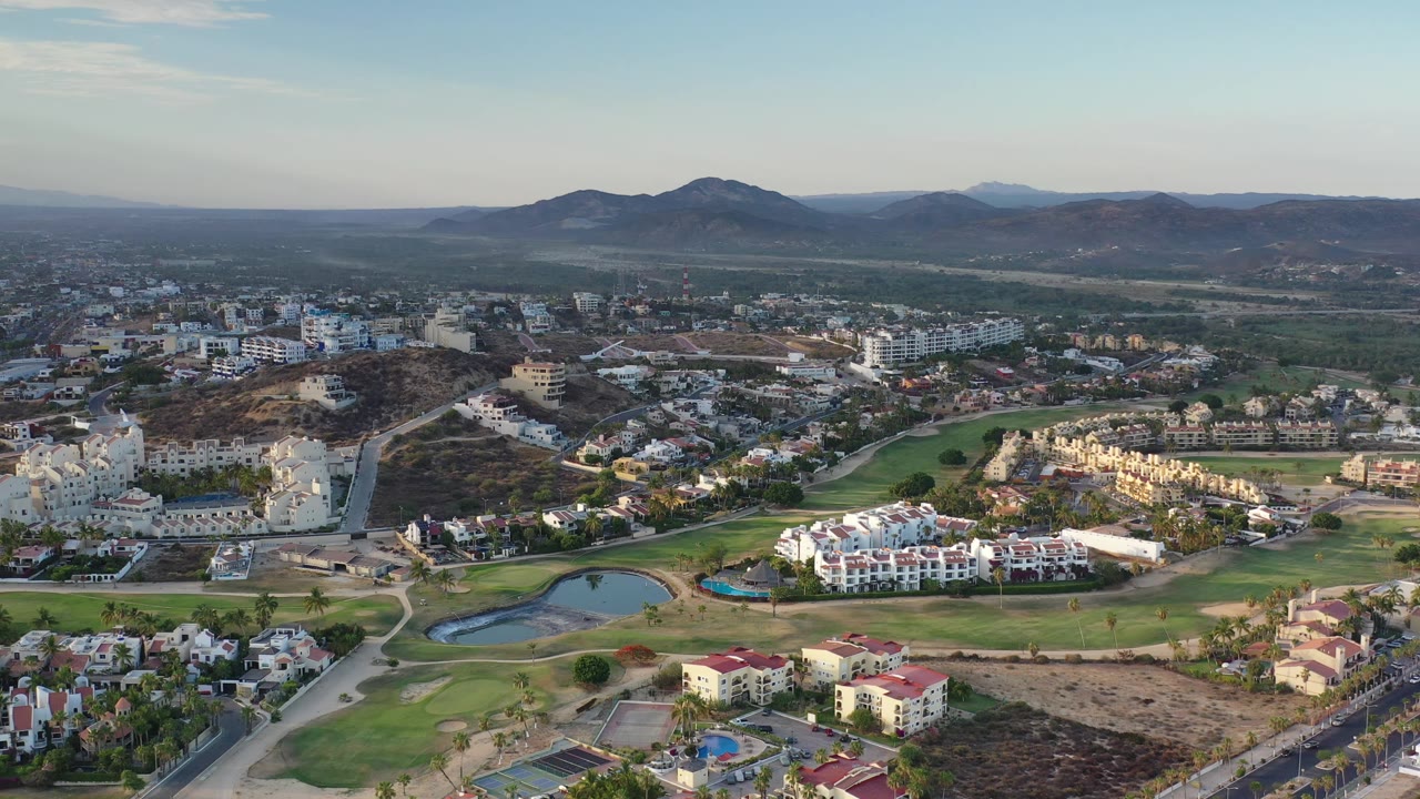 Aerial frame in Los Cabos, Mexico, with white apartment-style buildings, fairways, curving roads, ponds, dry terrain, and mountain ridges with no visible people.