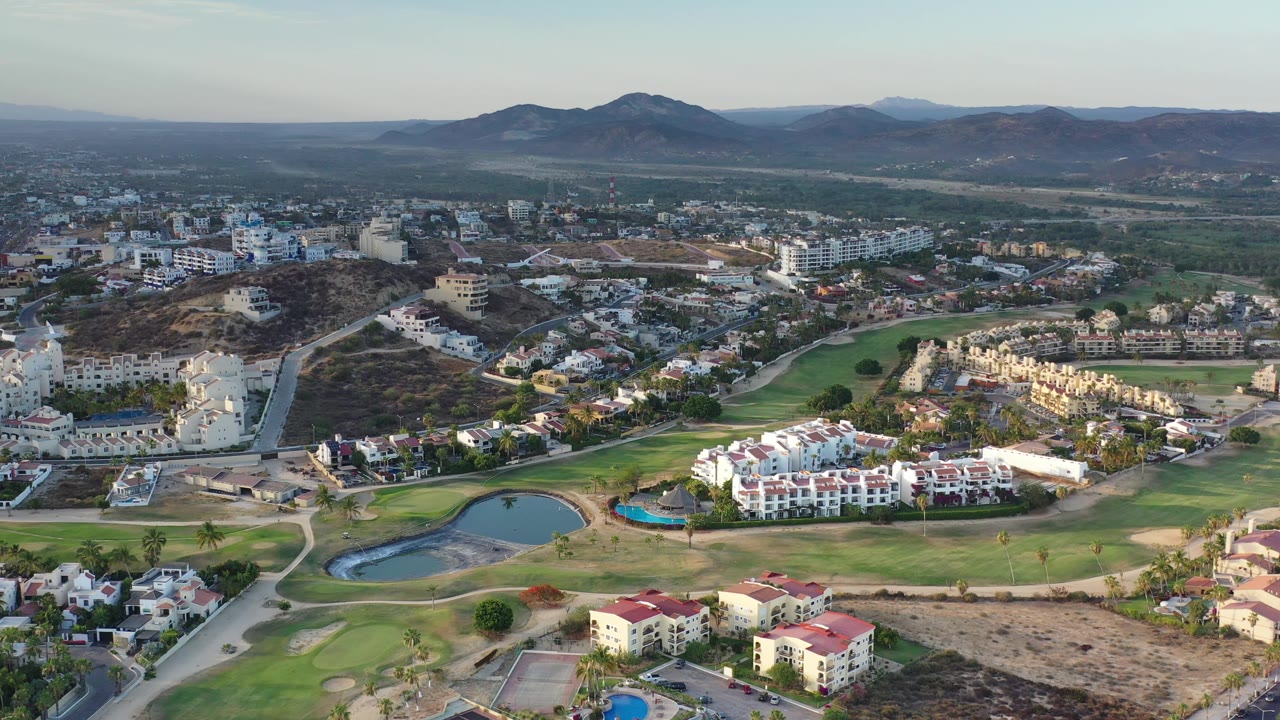 Los Cabos aerial view with the pond low in frame, resort-style buildings across the fairway, town blocks beyond, mountains on the horizon, and no visible people.