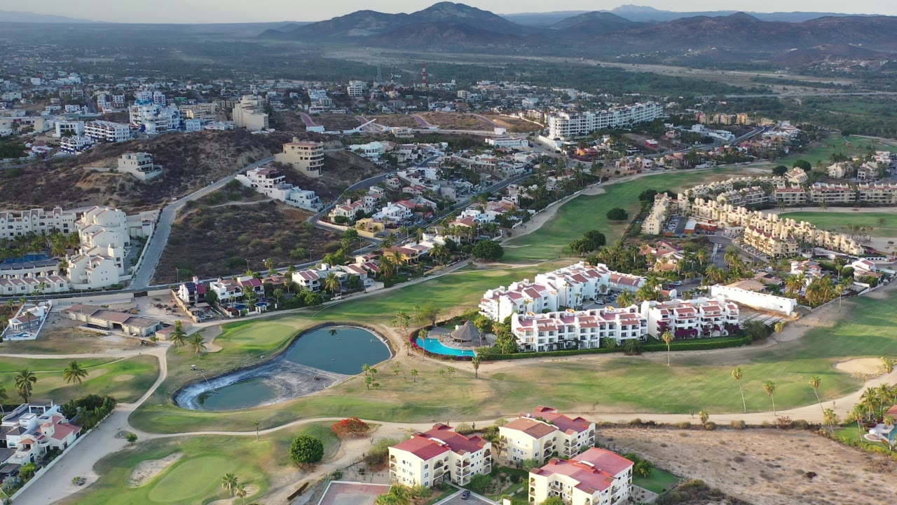 Aerial view over golf course greens, ponds, roads, homes, and distant hills.
