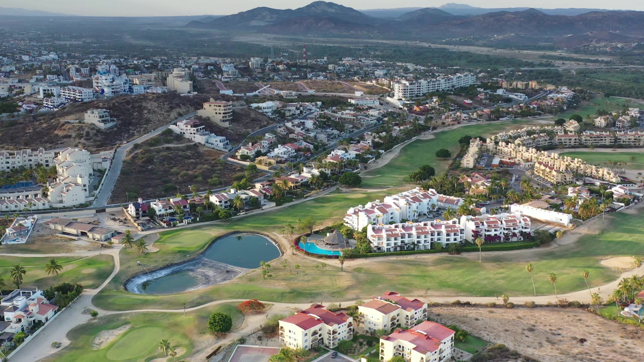 Aerial frame from Los Cabos, Mexico, with a rounded pond near the lower left, green fairways, pale roads, white villas, larger buildings, scrub-covered hills, and no visible people.