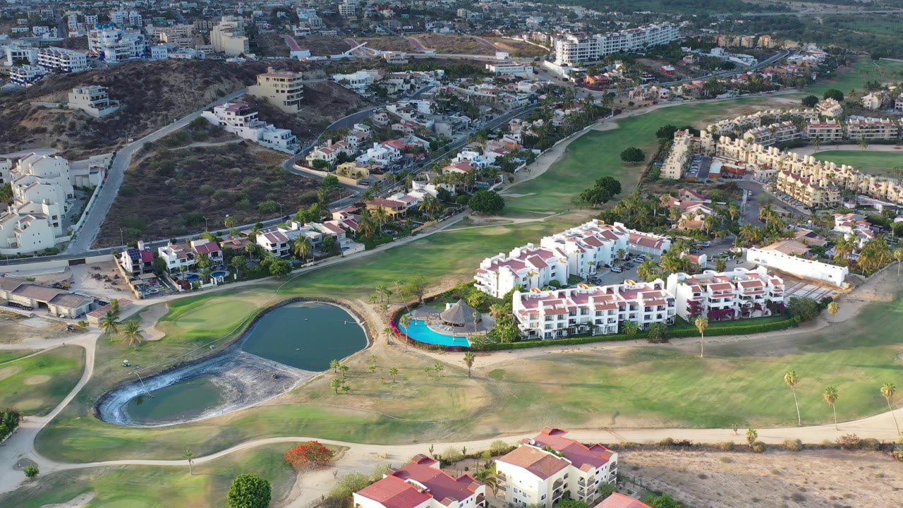 Aerial view in Los Cabos, Mexico showing a golf course pond, white residential buildings, red roofs, a blue pool, hillside roads, and no visible people.