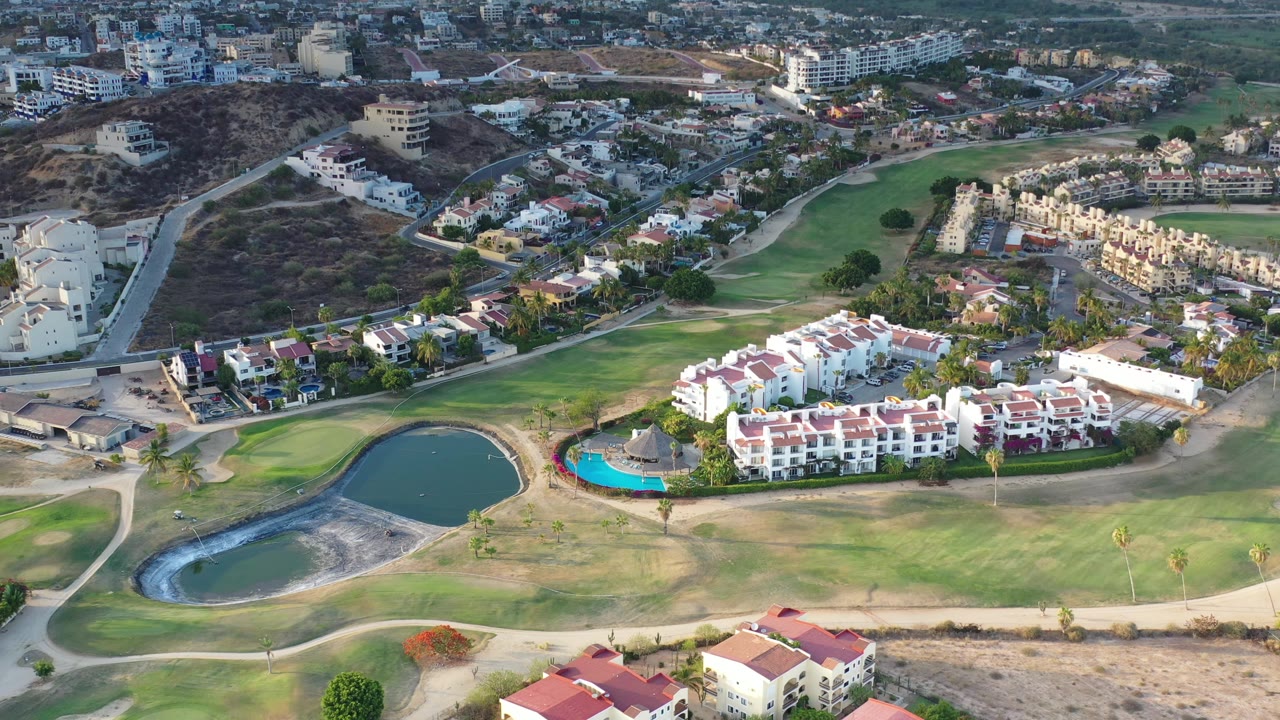 High aerial in Los Cabos, Mexico with the dark pond lower left, fairway grass across the center, white homes near a blue pool, and no visible people.