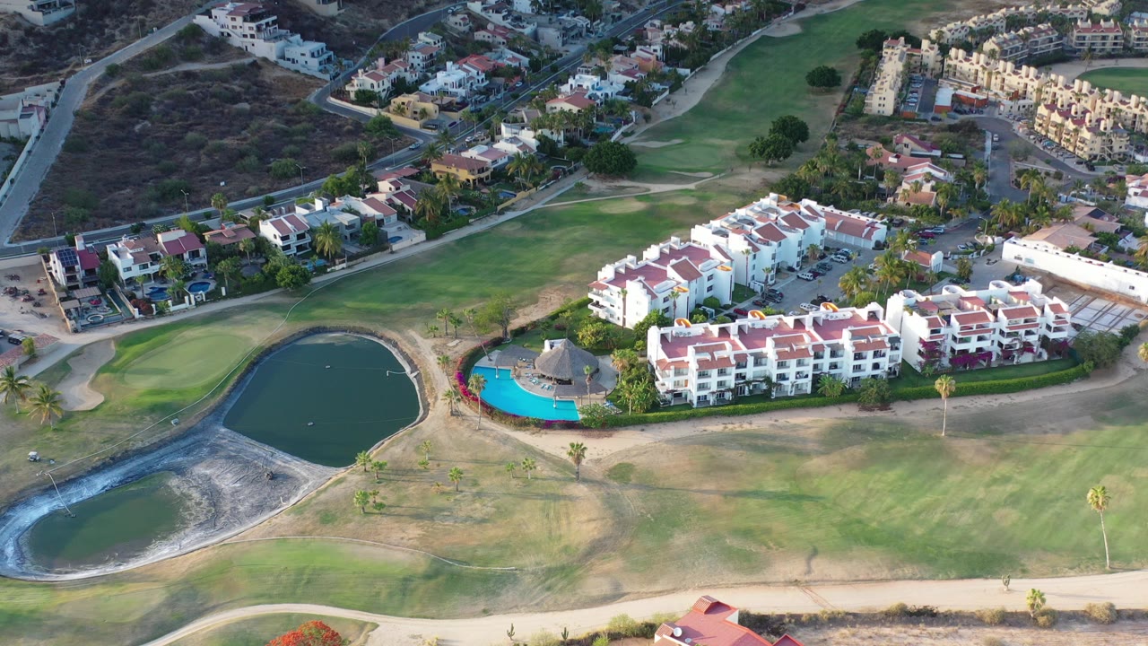 Elevated view over Los Cabos apartment buildings with red tile roofs, a bright blue pool, dark ponds, fairway grass, nearby roads, and no visible people.