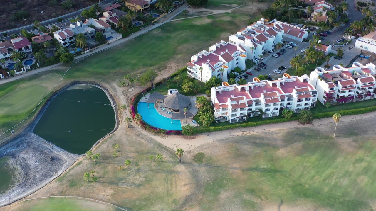 Aerial view of white resort buildings, red roofs, pool, golf course, and pond.