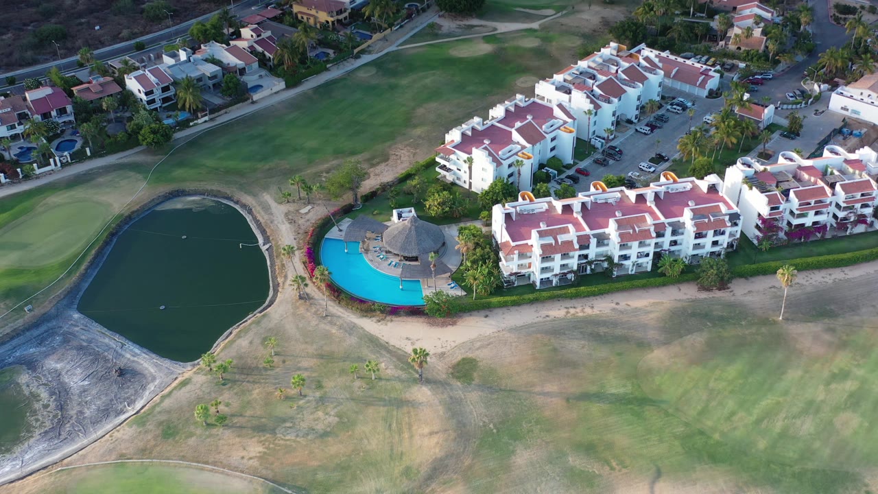 Los Cabos aerial frame with the pond on the left, a turquoise pool and palapa near the center, white resort buildings to the right, golf course grass, and no visible people.