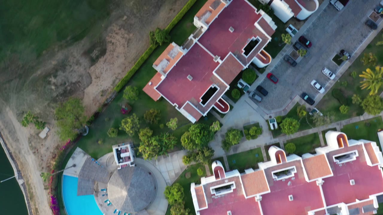Top-down Los Cabos frame with red and orange roofs, parked cars along the road, landscaped trees, a turquoise pool corner, and no visible people.