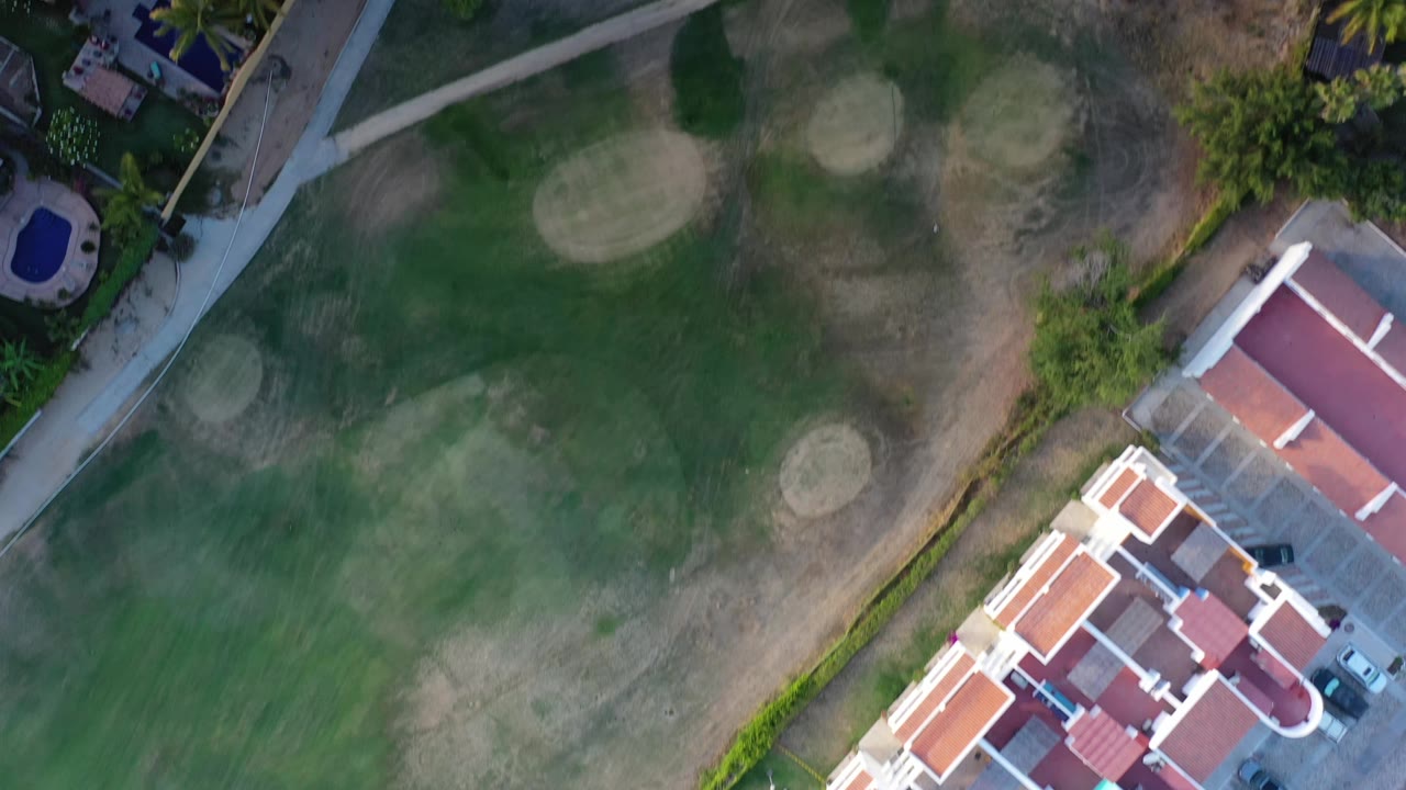 Los Cabos overhead frame with the golf course centered, dry patches across the fairway, red-roofed houses along the lower right, blue pools, palms, and no visible people.
