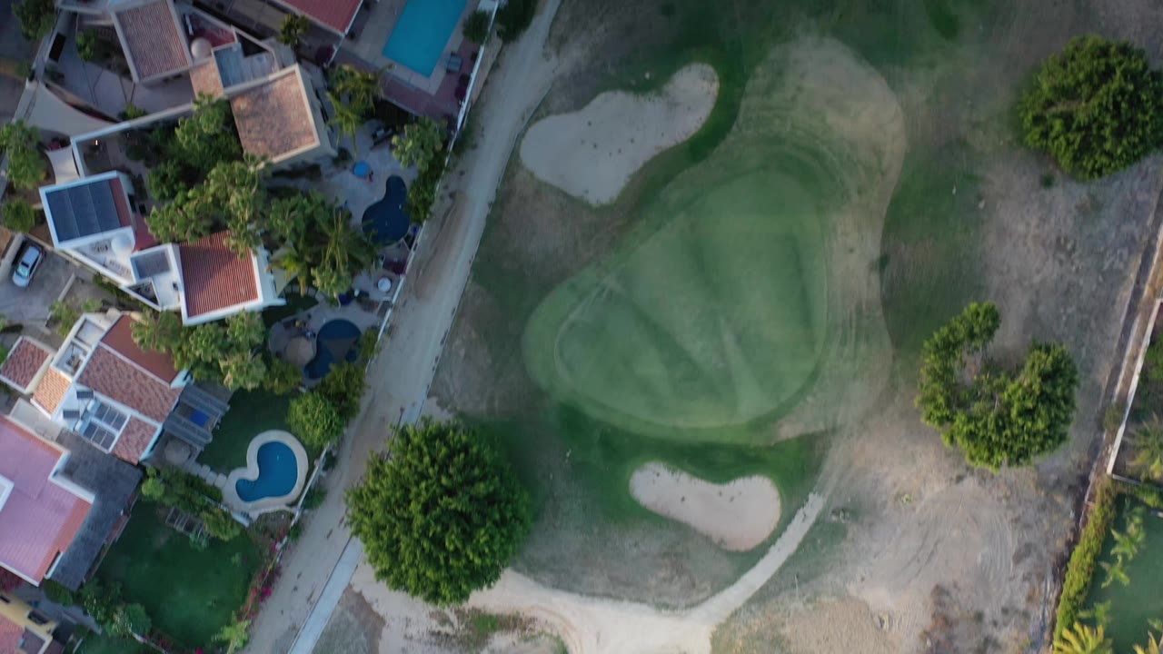 Overhead Los Cabos frame showing the putting green and pale sand traps centered beside a strip of homes, pools, palms, and dry fairway grass, with no visible people.