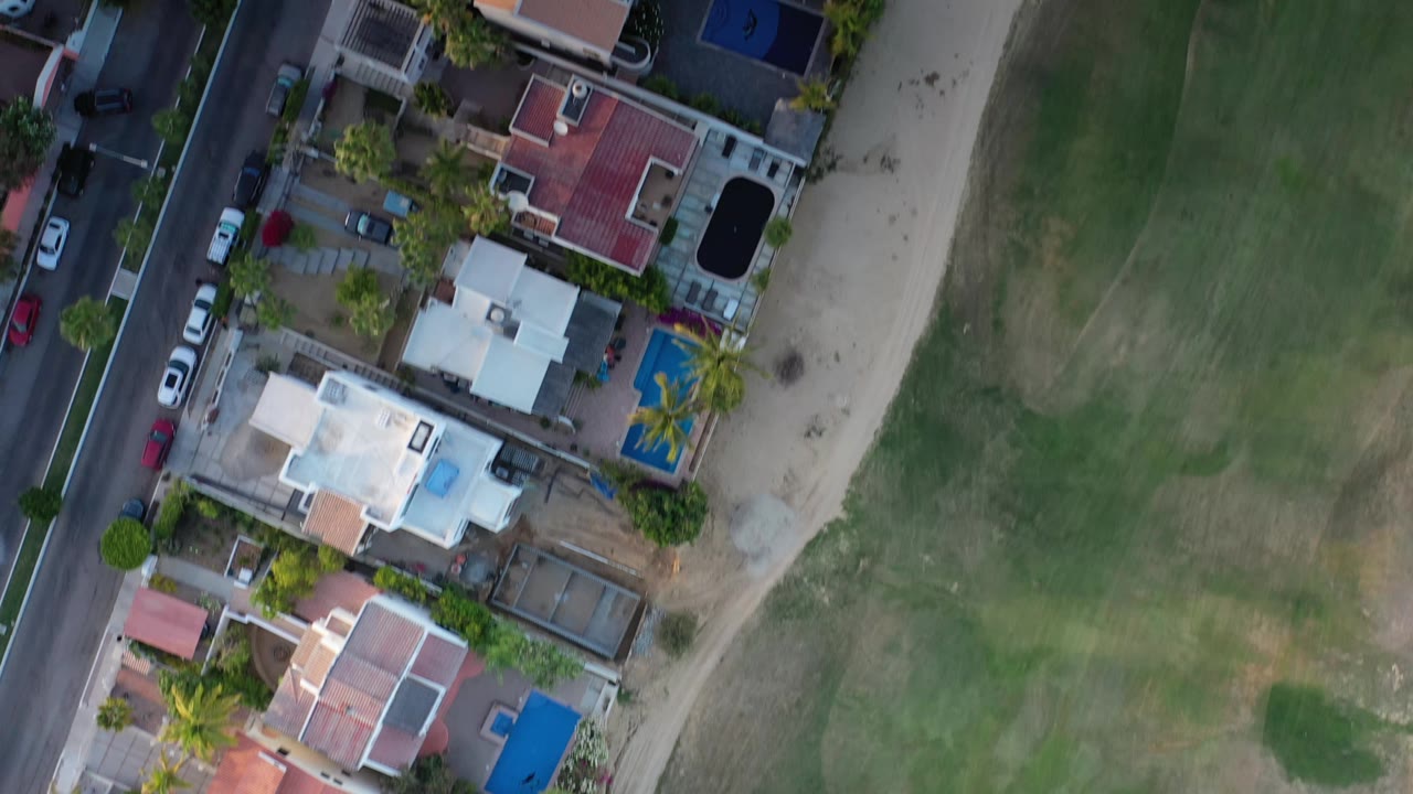 A later overhead view in Los Cabos keeps the road on the left and green field on the right, with several private homes, blue pools, parked cars, and no visible people.