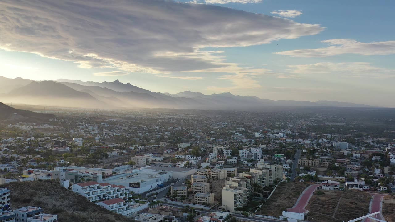 Los Cabos aerial frame with the sun peeking through the upper cloud edge, a spread of city buildings below, layered mountains on the horizon, and no visible people.
