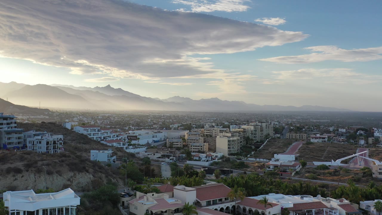 Opening aerial frame over Los Cabos, Mexico, showing hillside homes, low-rise city buildings, hazy mountains, a broad cloud bank, and no visible people.