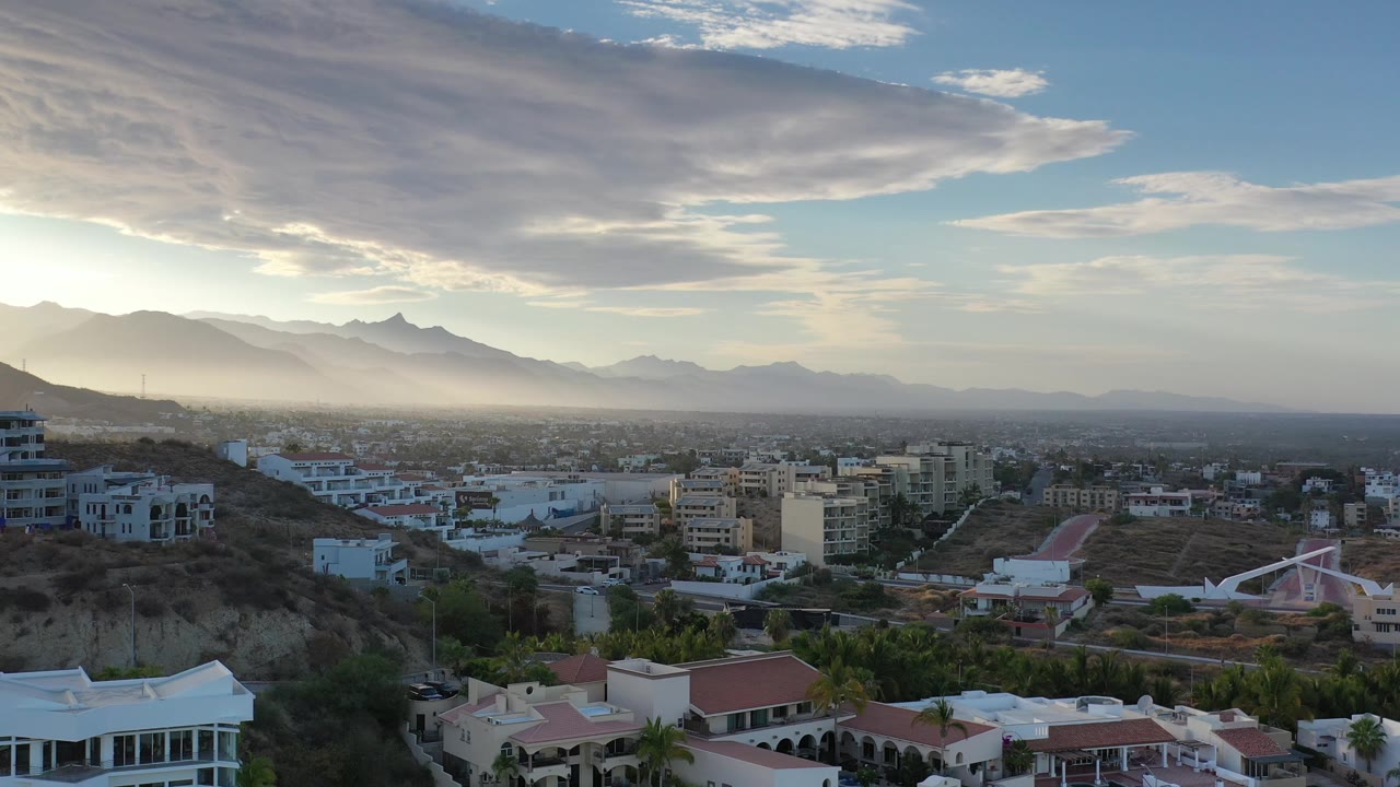 Aerial view of Los Cabos rooftops, hillside terrain, mountains, and layered afternoon clouds.