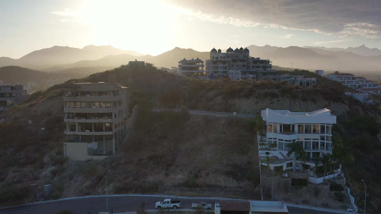 Los Cabos appears from an elevated angle with white hillside homes, a dry slope, a bright sun near the mountain ridge, and no visible people.